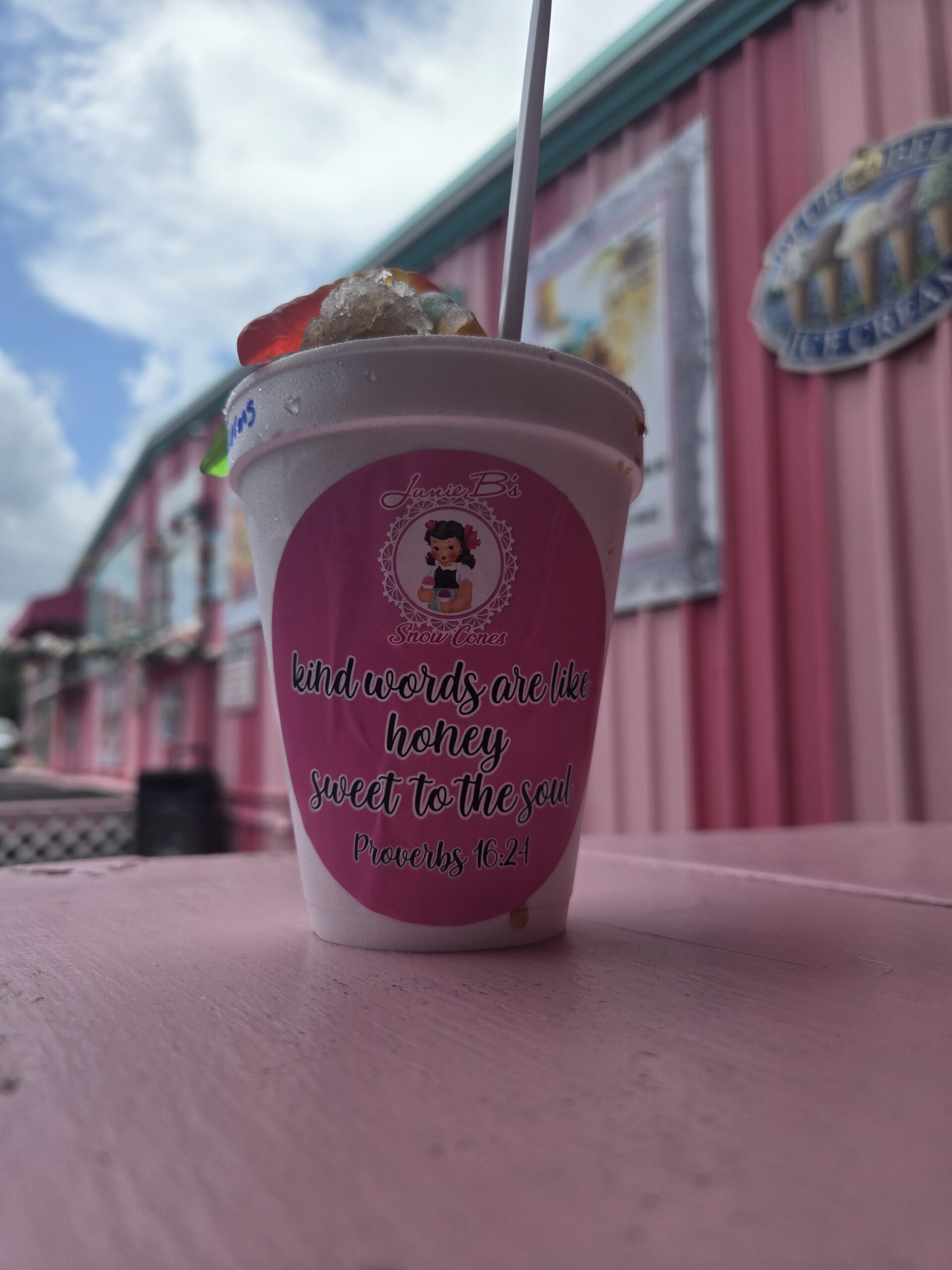 A colorful snowcone topped with gummy worms sitting on a pink table outside Junie B’s Snowcones storefront.