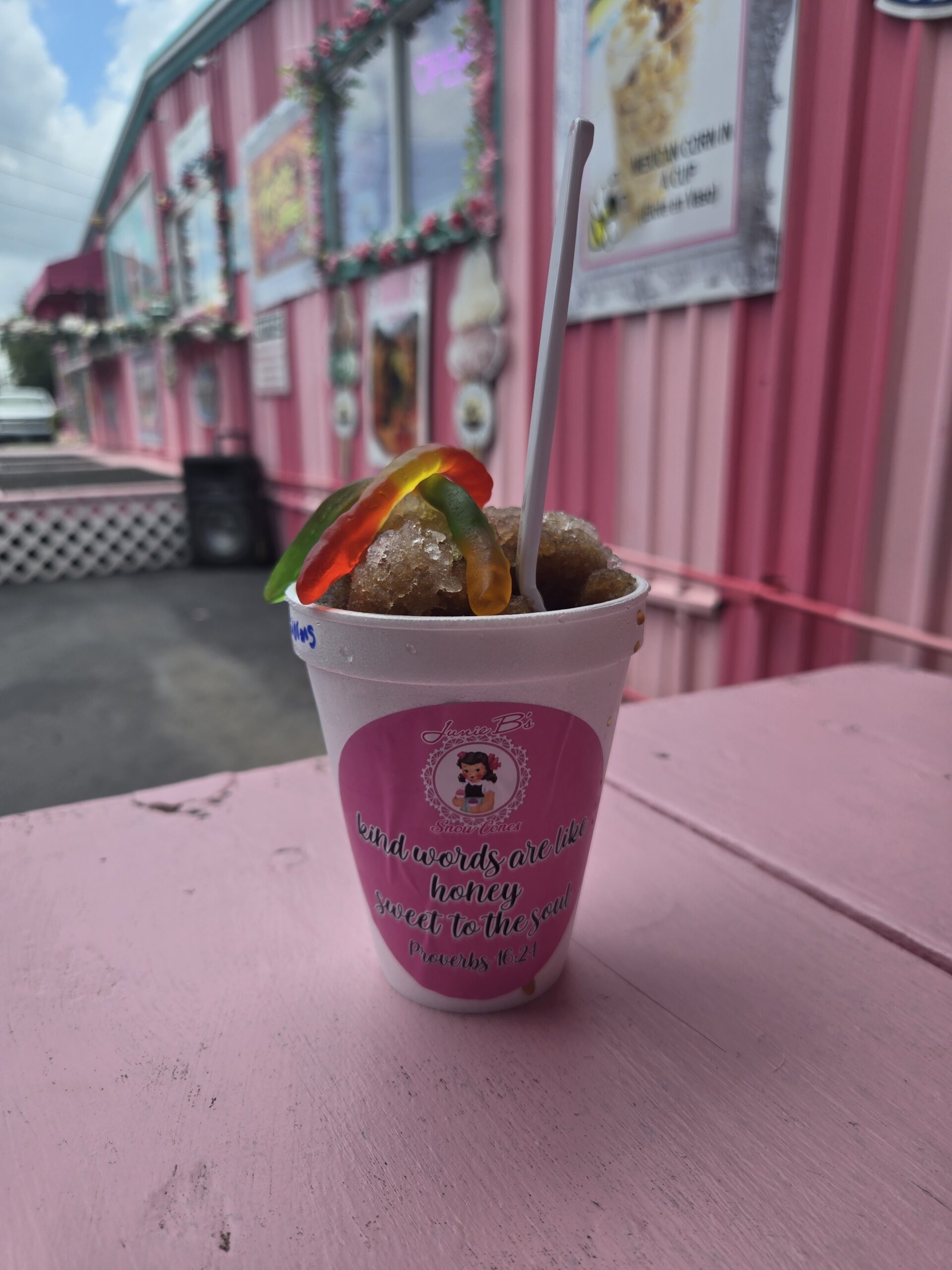 A colorful snowcone topped with gummy worms sitting on a pink table outside Junie B’s Snowcones storefront.